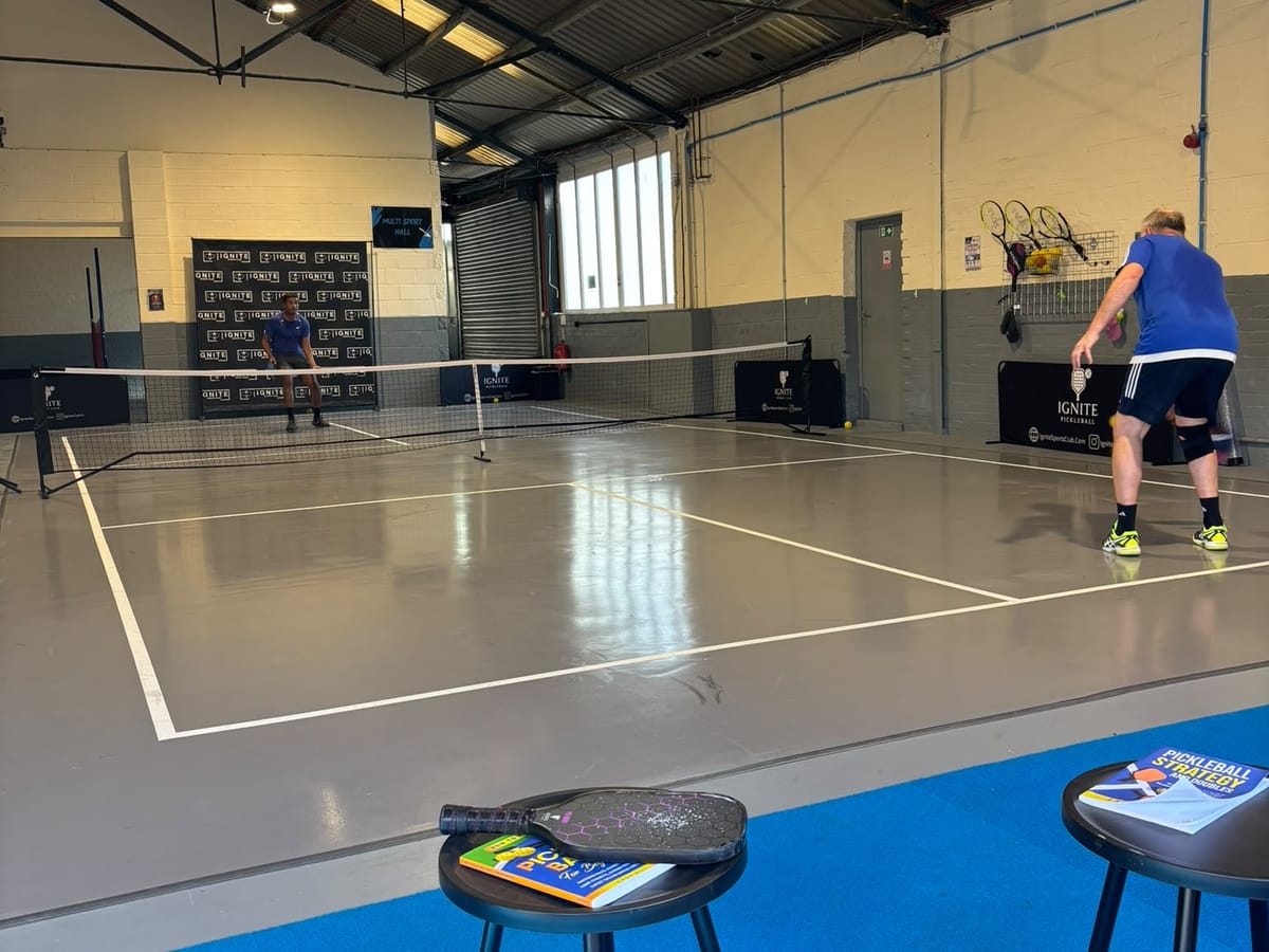A daytime pickleball rally on the grey court at Ignite, with rackets hung on the wall behind