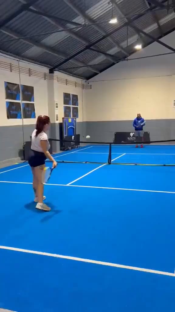 Two players in a rally on the blue indoor court at Ignite Sports Club at night, under the hall lights